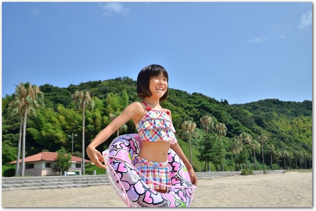 海辺にフリルのついた水着を着て浮き輪を持った女の子が立っている光景